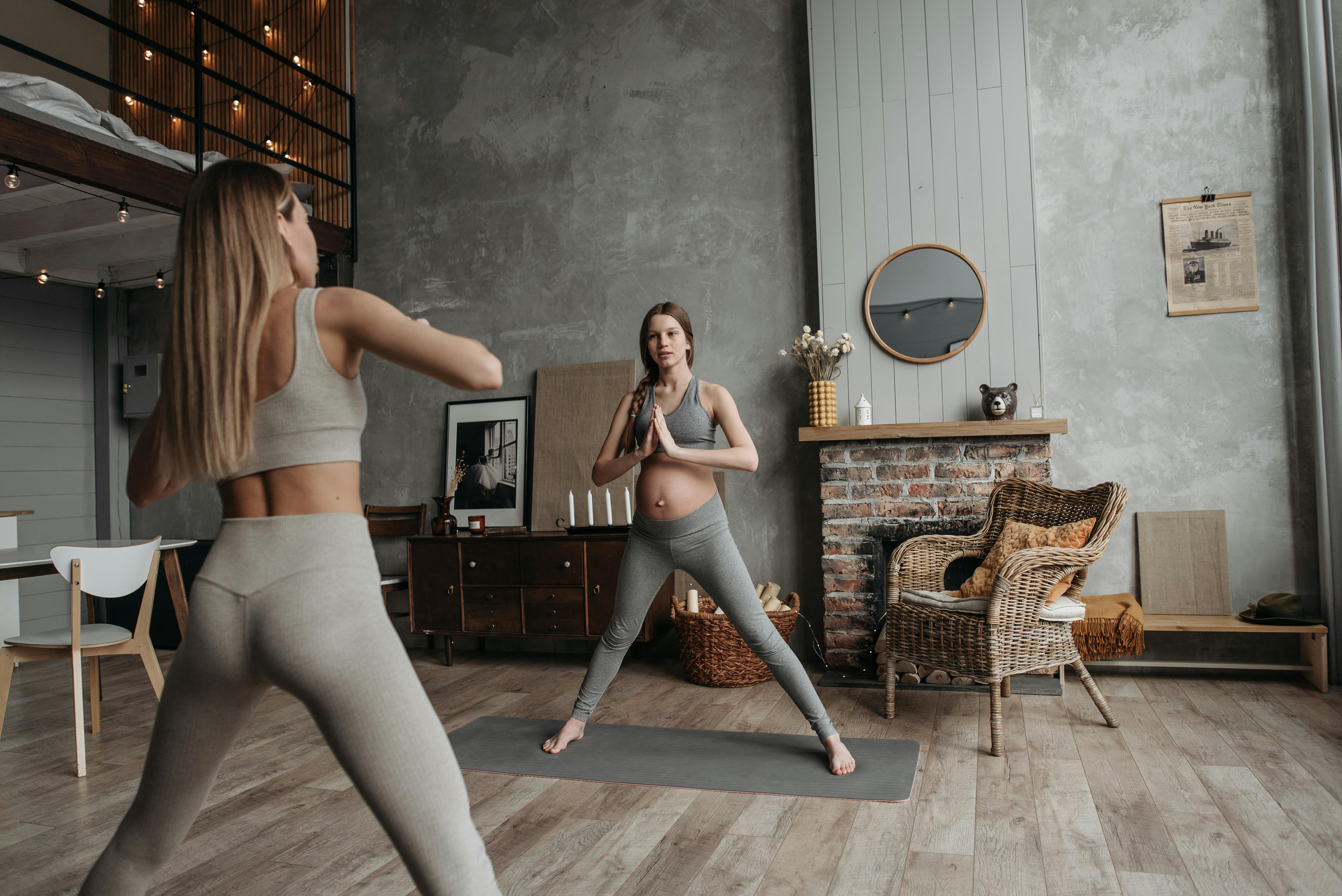A pregnant woman in activewear performing yoga poses with a trainer in a stylish indoor setting.
