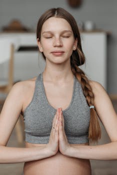 Pregnant woman meditating with eyes closed and hands in Namaste pose, wearing a sports bra.