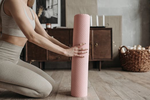 A woman in activewear unrolls a pink yoga mat indoors on a wooden floor.