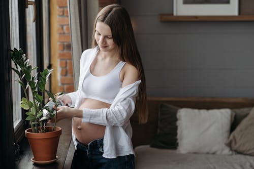 Woman in White Crop Top and Blue Denim Jeans Watering a Potted Plant