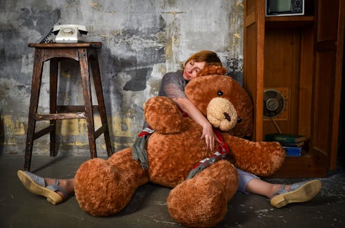 Full length young female hugging big plush toy bear and sitting with eyes closed and legs spread on floor