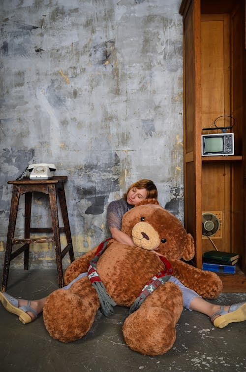 Serene young female embracing big plush bear and sitting on floor with eyes closed and legs spread