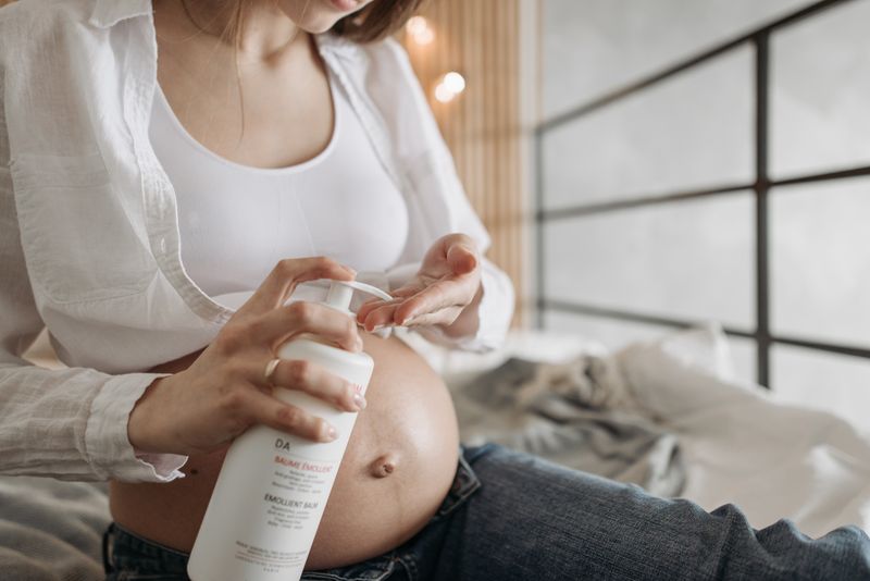 A pregnant woman applying lotion to her belly while sitting indoors wearing casual clothes.