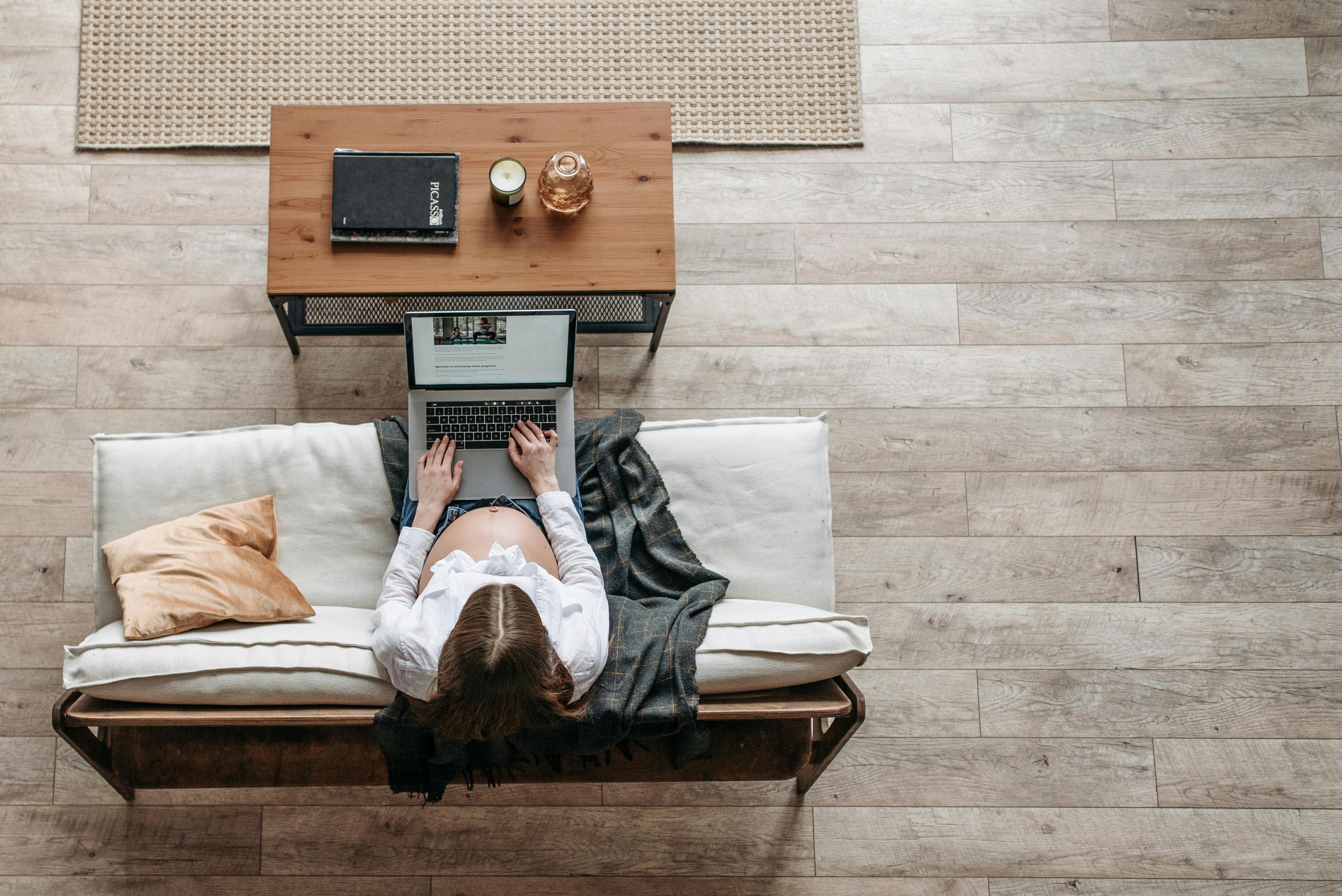 High-angle view of pregnant woman on sofa using a laptop in a warm living room setting.