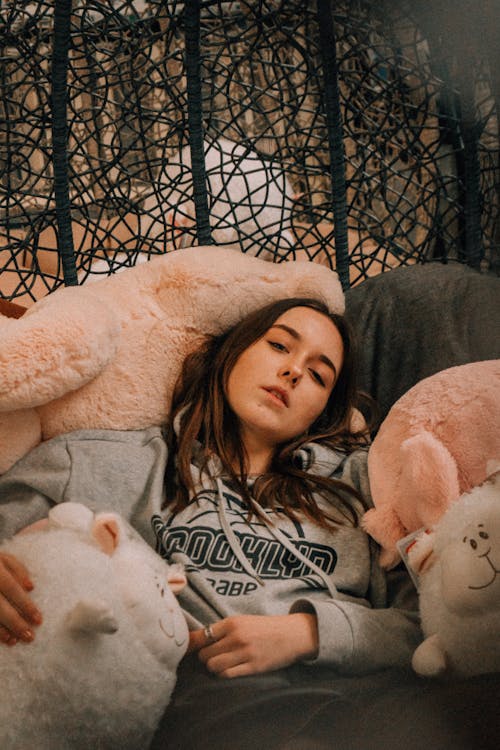 Calm young female in hoodie looking at camera while lying amidst soft toys near black metal fence with decorative elements