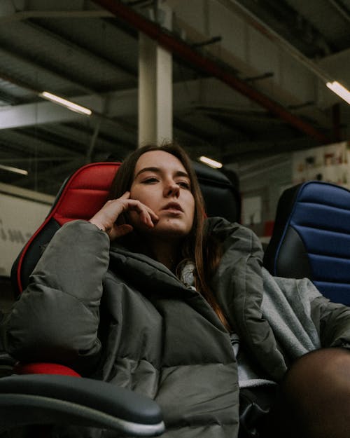 Focused female in warm clothes looking away while sitting in comfortable armchair near glass window in modern stadium during game