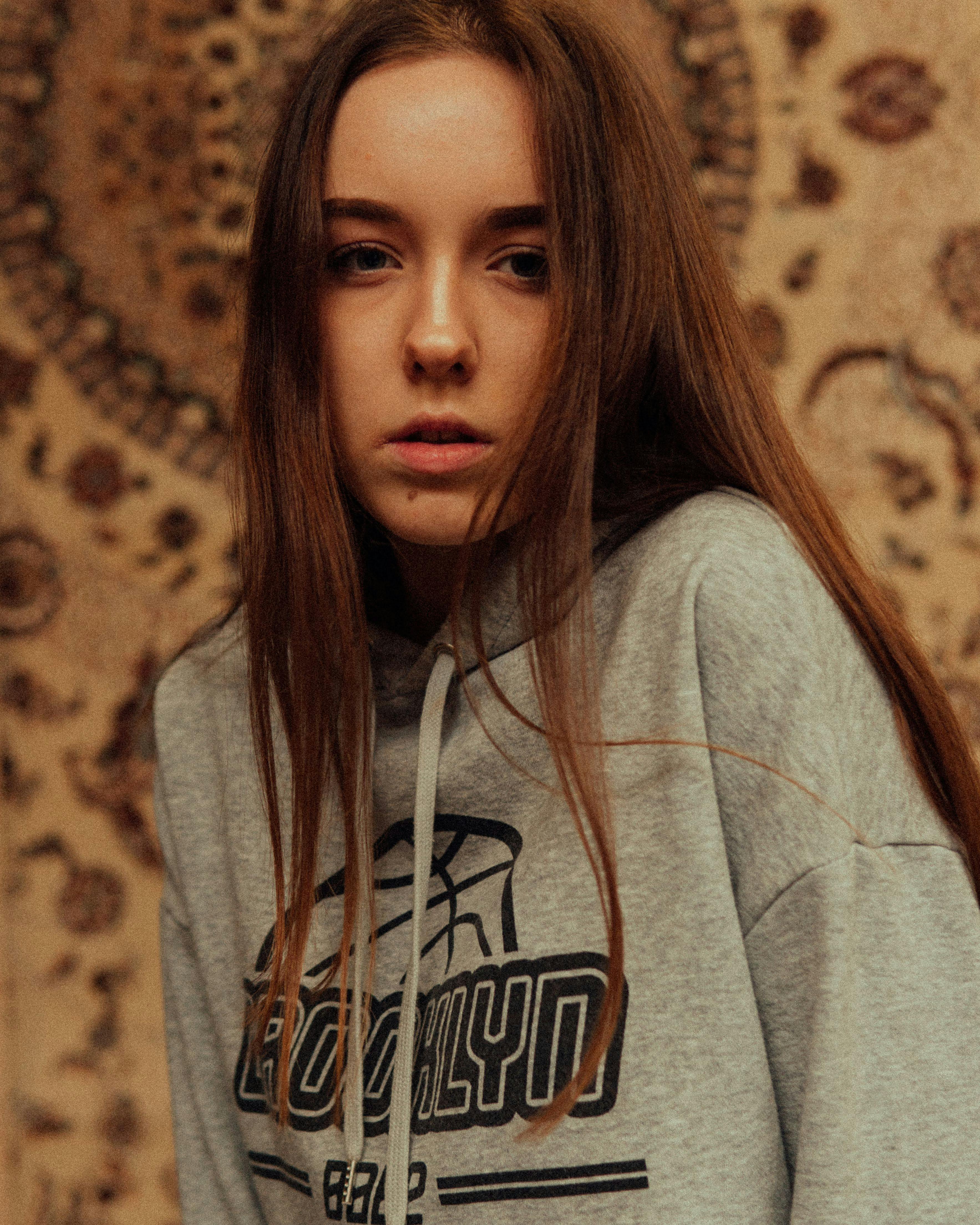 Serious female in casual gray wear with long hair looking at camera while standing near wall with old fashioned carpet
