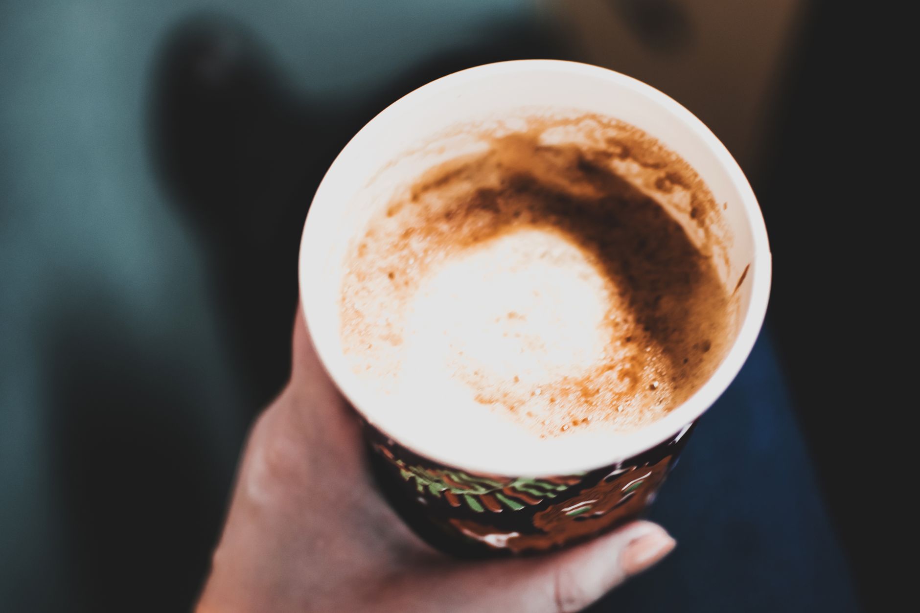 Close-up view of a hand holding a hot cappuccino with frothy foam in a decorative paper cup.