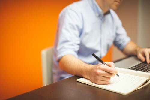 Focus Photography of Person Writing on Desk Using Laptop