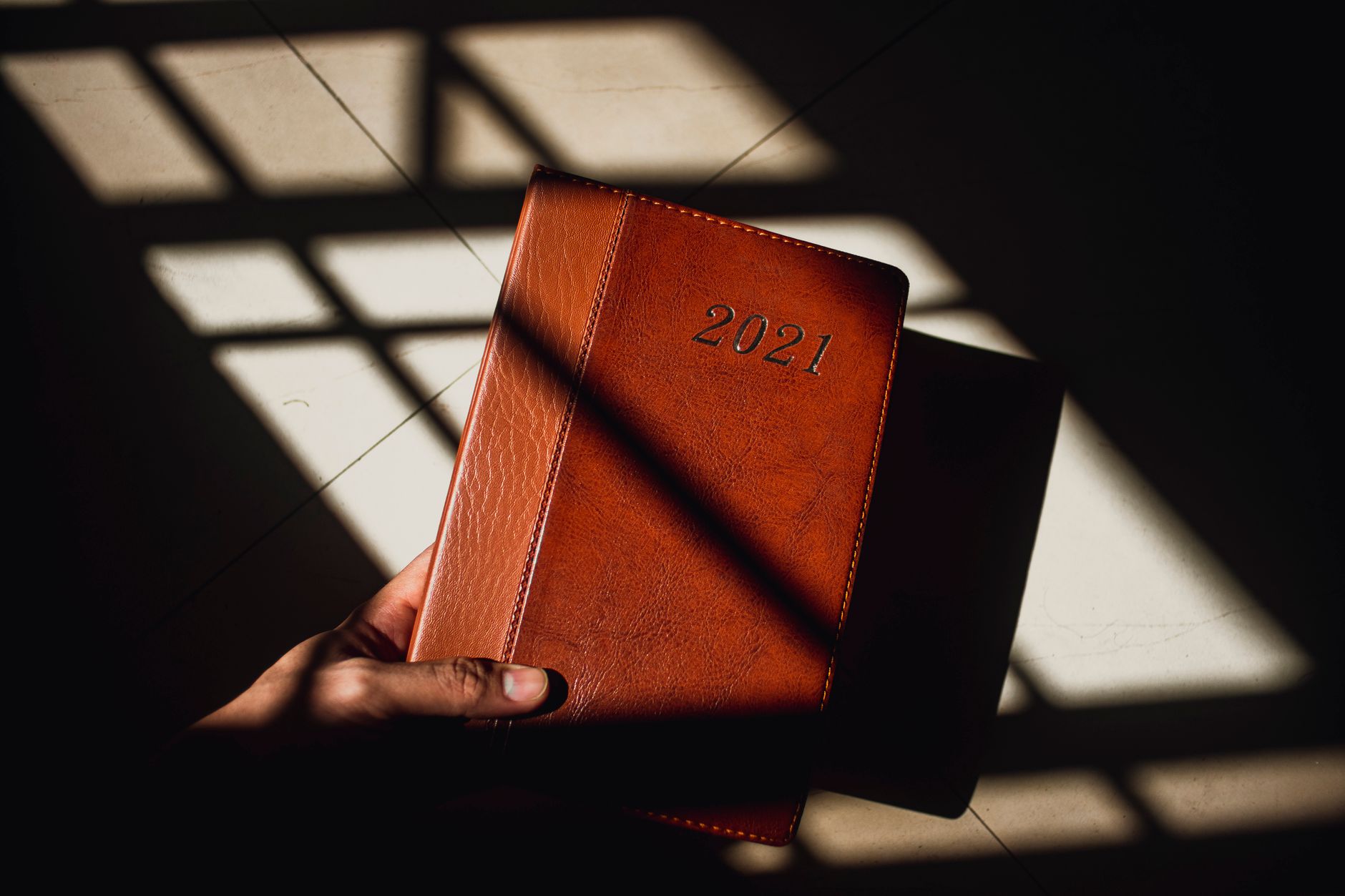 A hand holds a brown leather planner with '2021' embossed, illuminated by sunlight through a window grid.