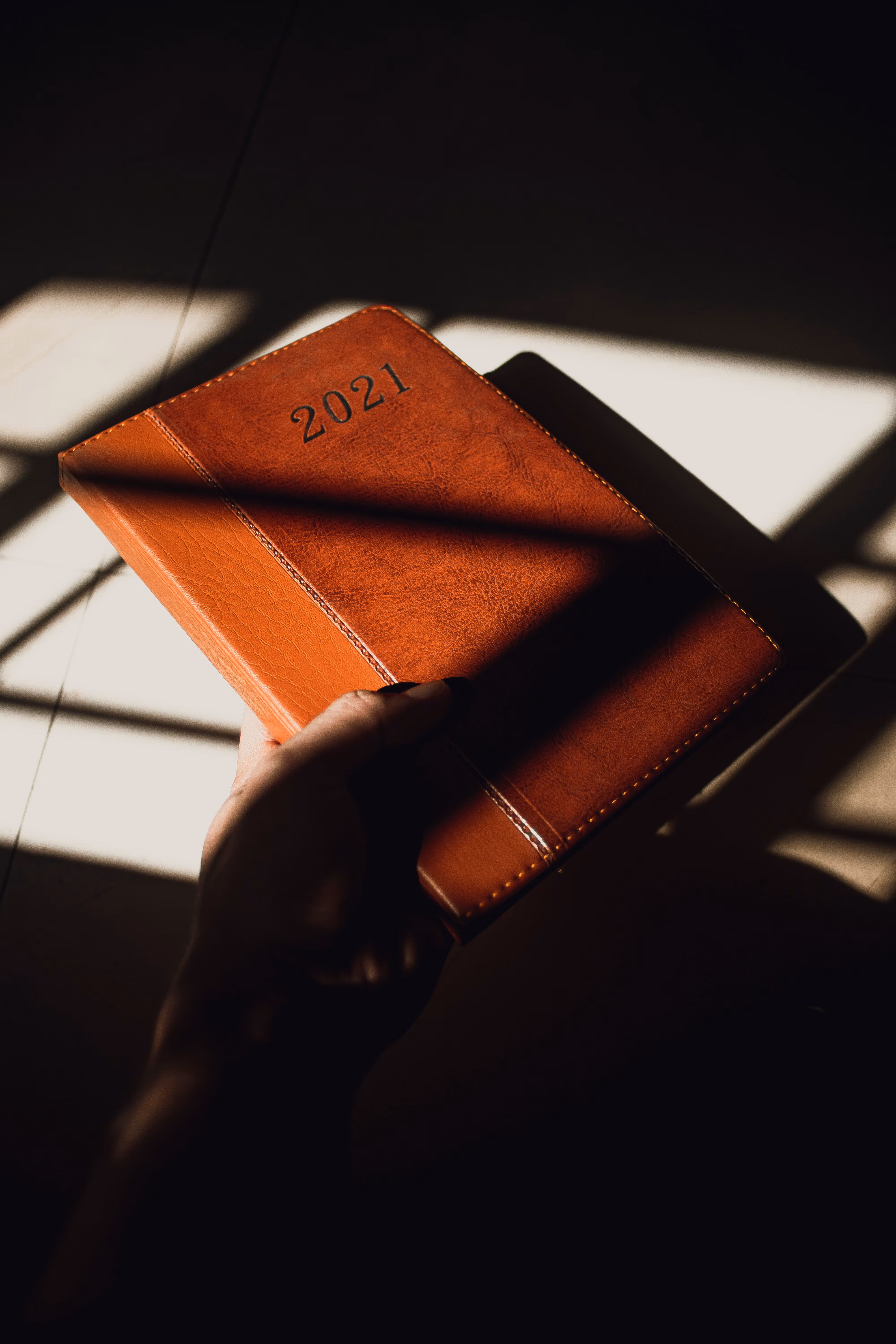 A hand holding a brown leather planner marked 2021 in dramatic shadows.