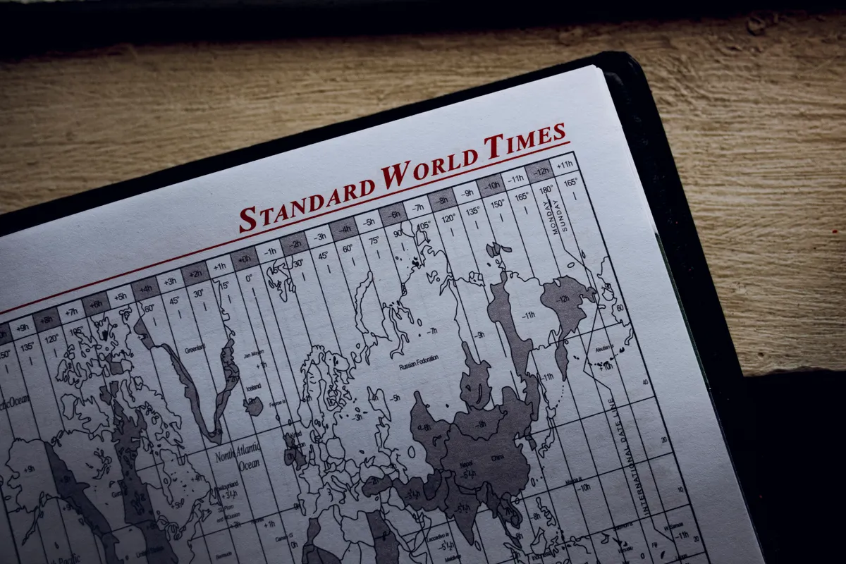 A printed Standard World Times map in a notebook on a wooden surface, showing continents overlaid with a grid of hour-offset columns.