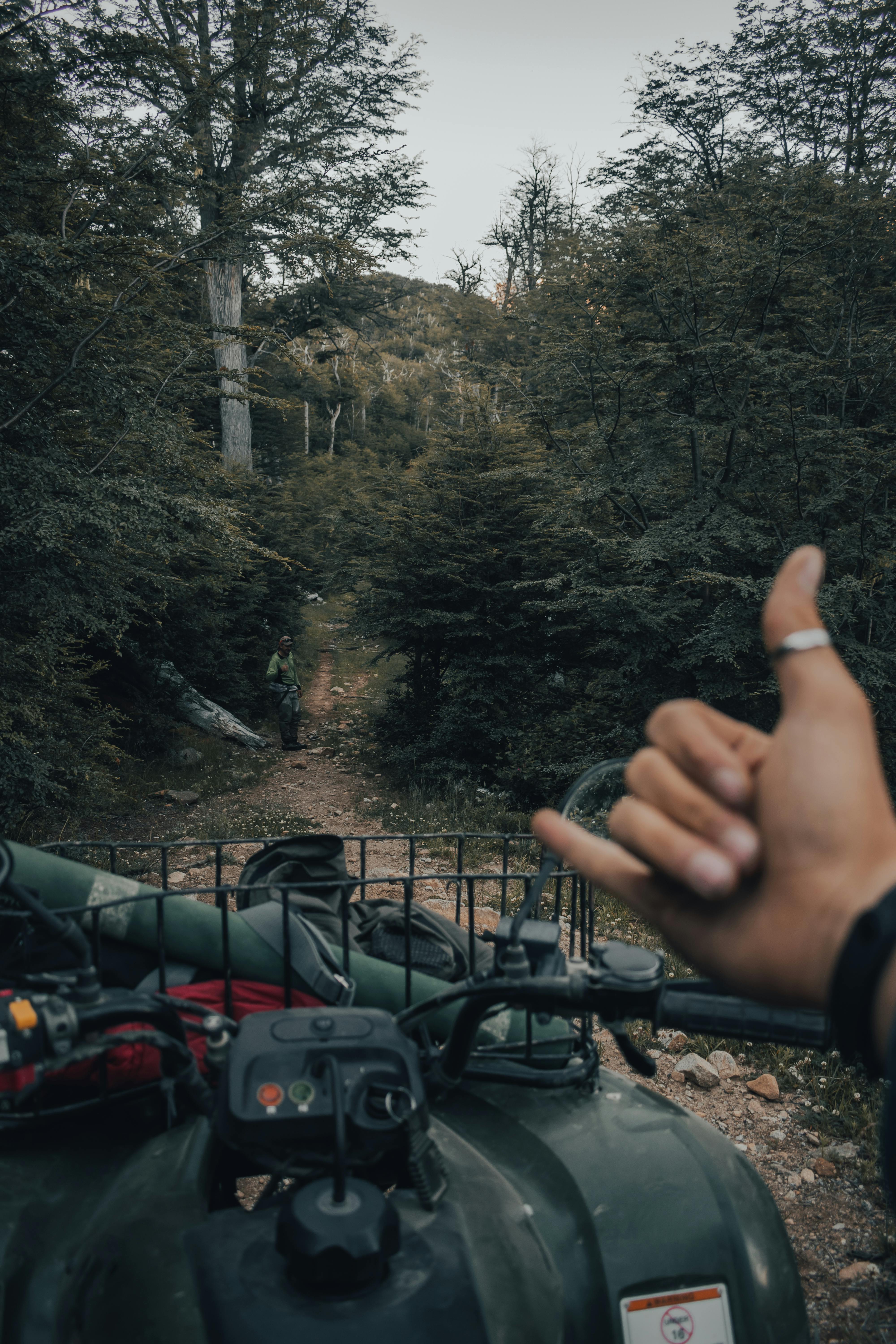 POV shot of hand signaling hang loose over motorbike in a forest path.
