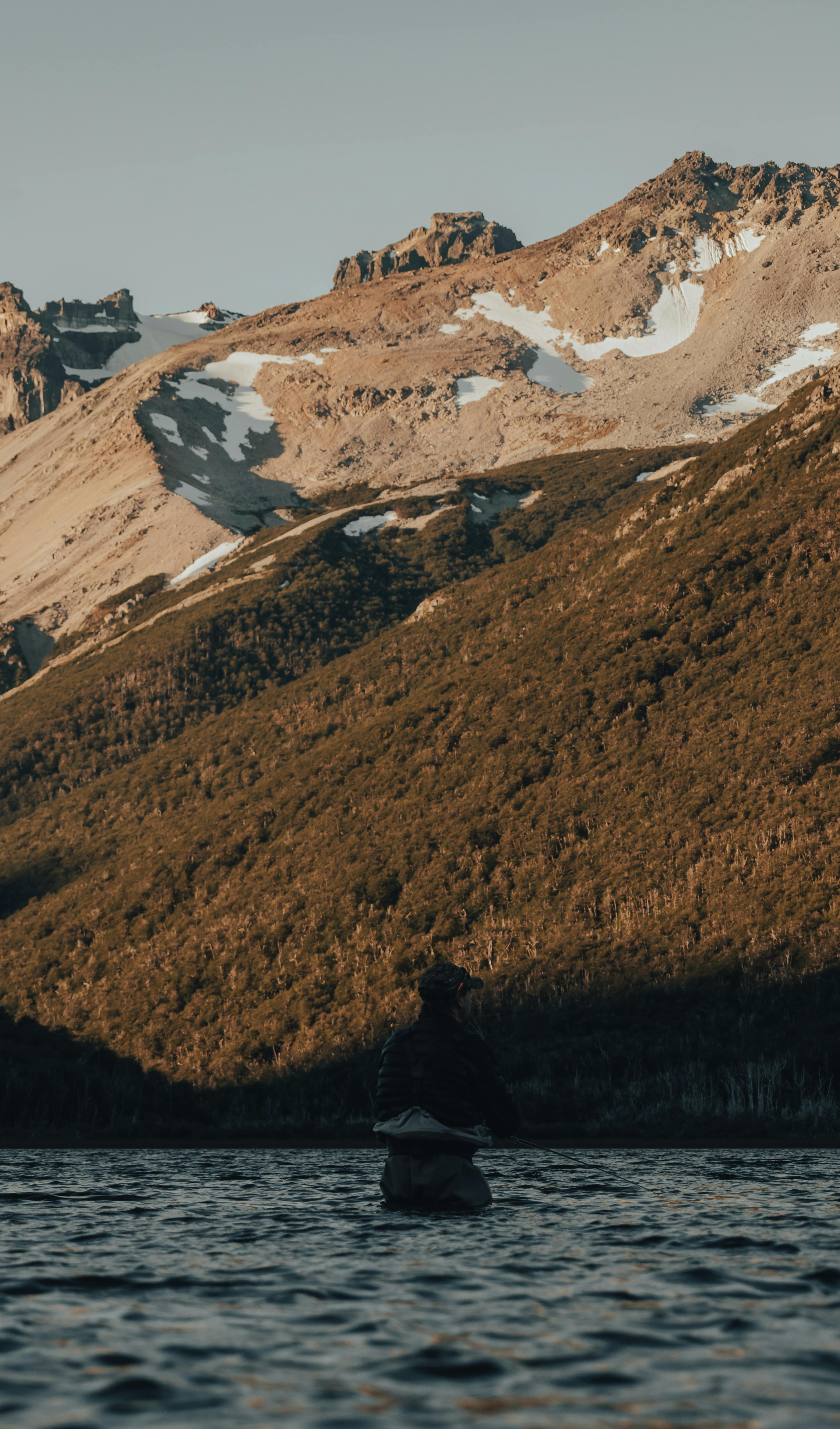 Person fishing in a tranquil lake against majestic mountain backdrop at sunrise.