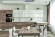 Modern open plan kitchen with white glossy and wooden cabinets and dining table with white chairs near in light apartment