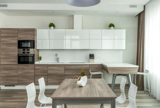 Modern open plan kitchen with white glossy and wooden cabinets and dining table with white chairs near in light apartment