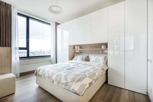 Interior of modern bedroom with white built in wall wardrobe and shelf above cozy bed with pillows and soft blanket in daylight