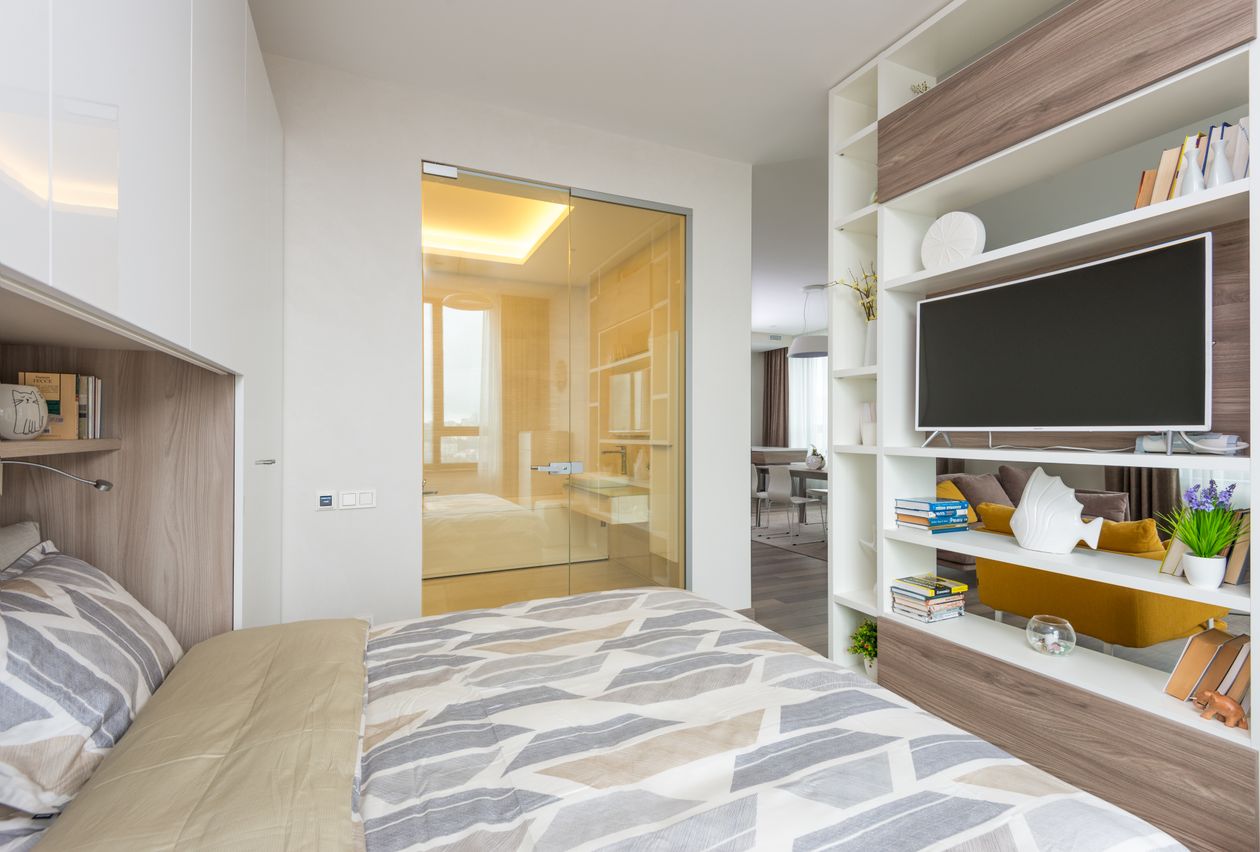 A white cube shelf with a TV mounted in the center, dividing a small sitting area from a bed