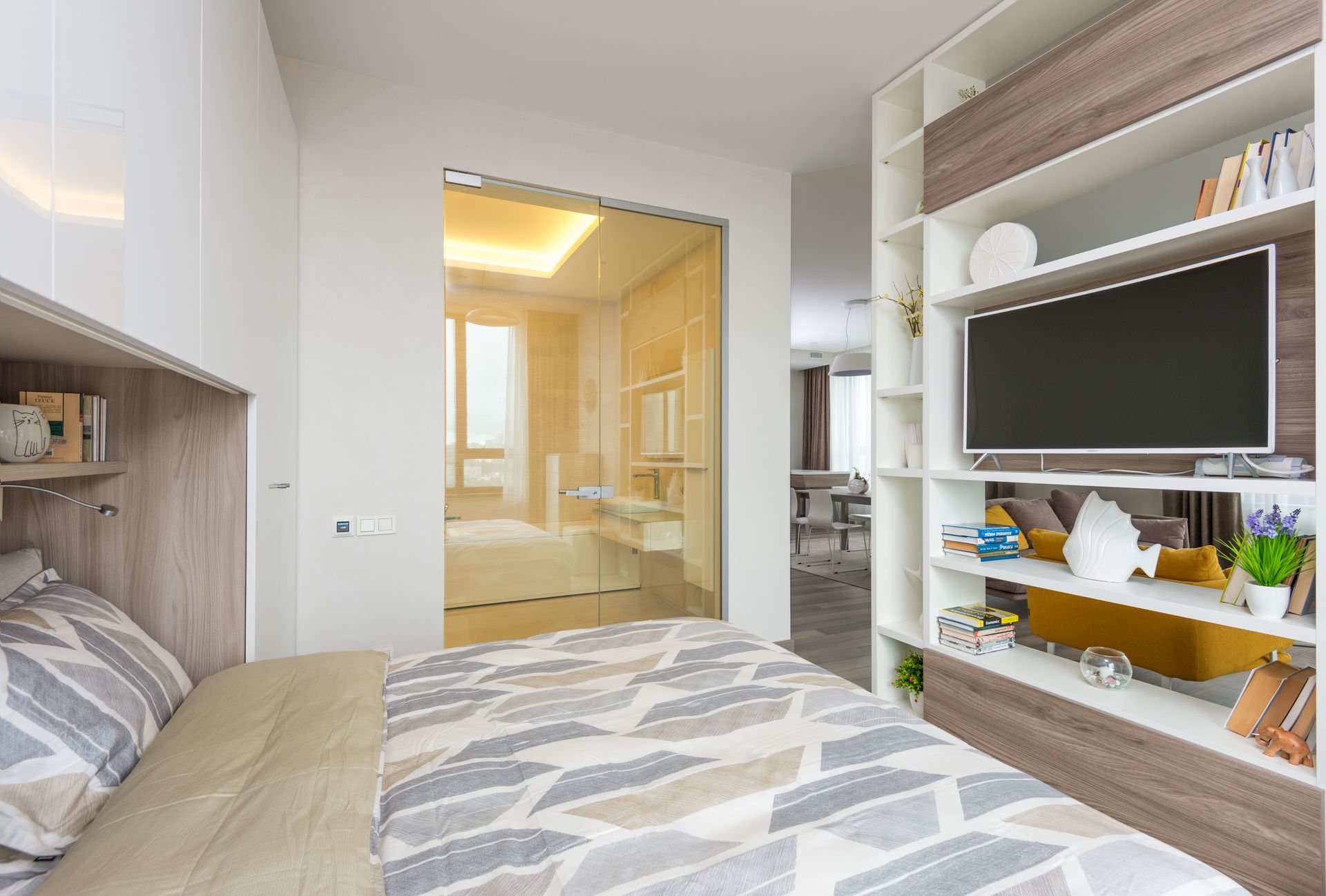 Interior of light cozy bedroom with open plan living room behind shelf partition with TV and opened glass doors to bathroom