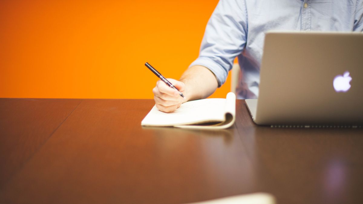 Adult writing notes in a notebook beside a laptop on a wooden desk with orange background.