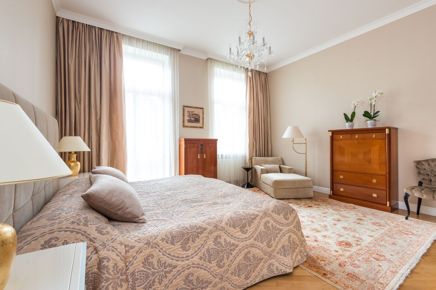 Pale bedroom with a crystal chandelier, upholstered bed, old wood dresser, floor lamp and patterned rug