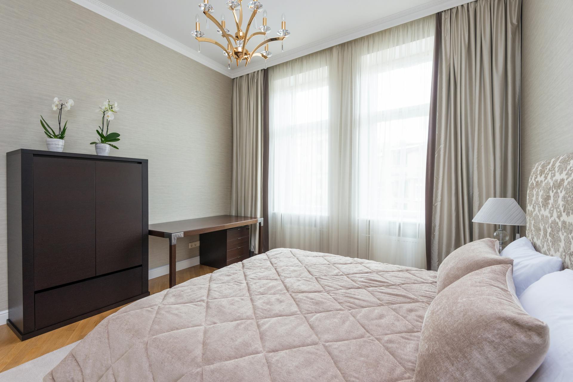 Interior of bedroom with coverlet and soft cushions placed near windows and wooden cupboard decorated with orchids