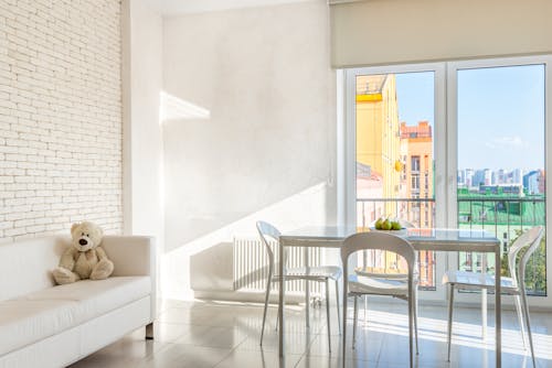 Empty white dining room with plush bear on sofa near dining table placed on tiled floor near panoramic balcony doors