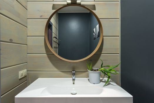 Round mirror hanging above sink with faucet and potted plant on wall made of panels in modern bathroom