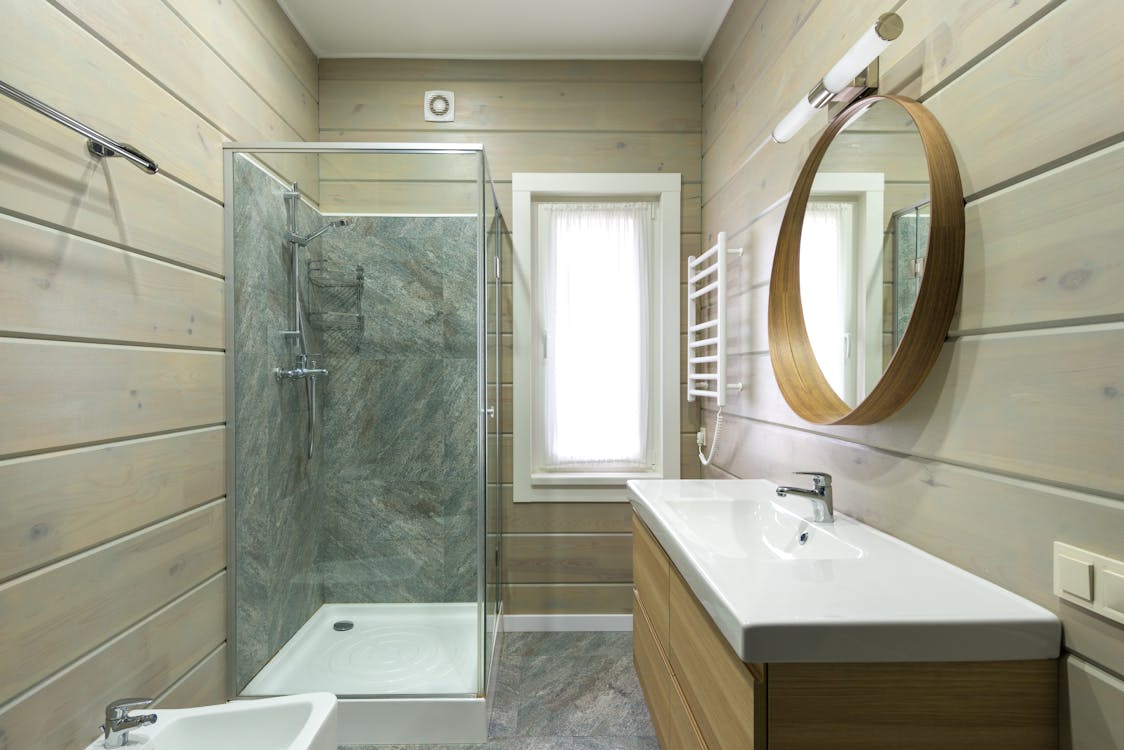 Free Contemporary bathroom design with round mirror hanging above sink and shower cabin near window and paneled walls Stock Photo