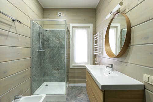 Contemporary bathroom design with round mirror hanging above sink and shower cabin near window and paneled walls