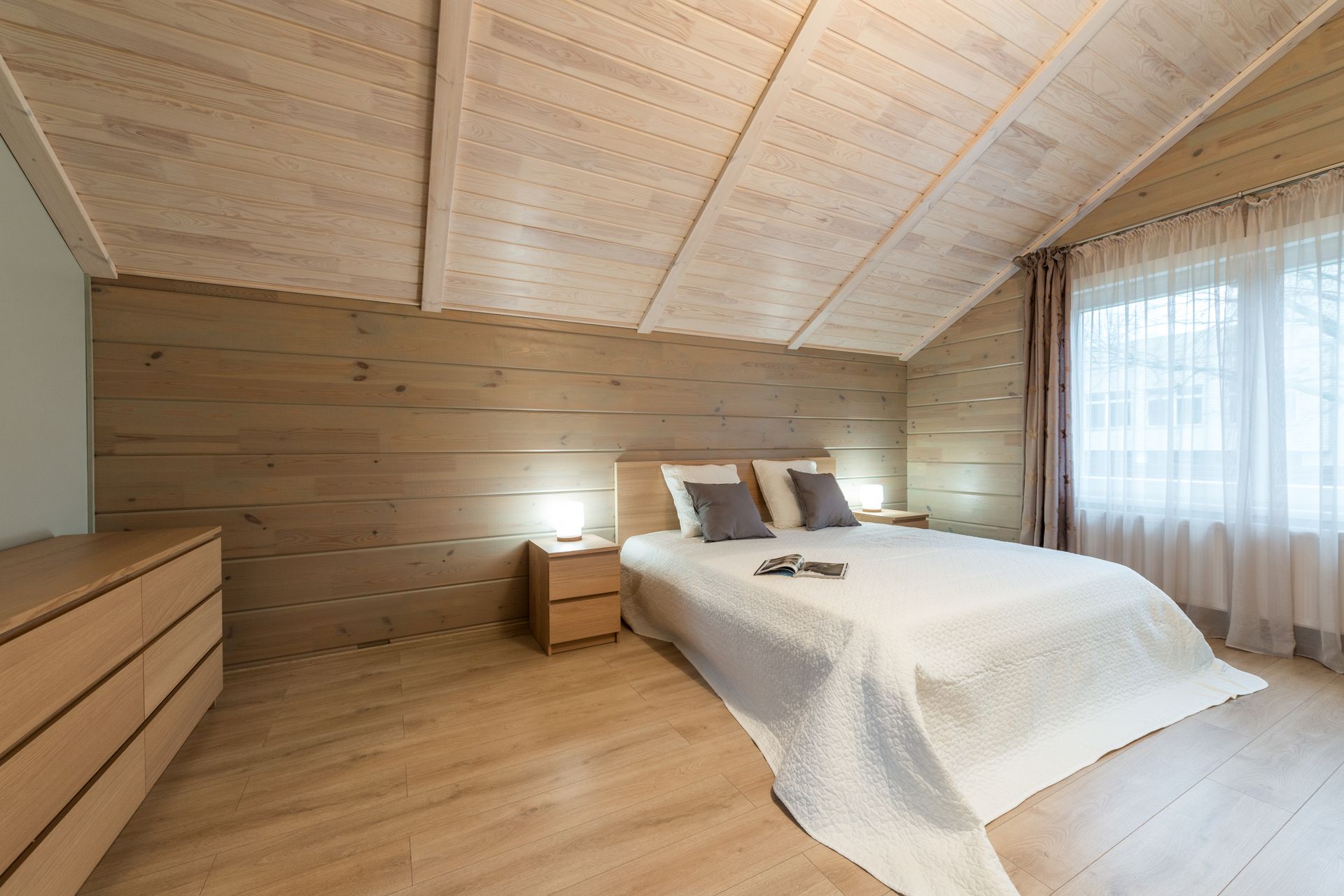 Comfortable bed with pillows and opened magazine with luminous lamps on nightstands on wooden mansard of cottage