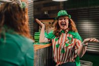 Cheerful adult female with curly red hair yelling and shrugging while chatting with unrecognizable friend in bar during Saint Patricks Day