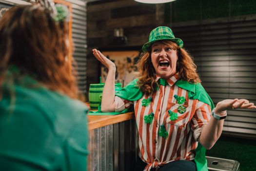 Cheerful adult female with curly red hair yelling and shrugging while chatting with unrecognizable friend in bar during Saint Patricks Day