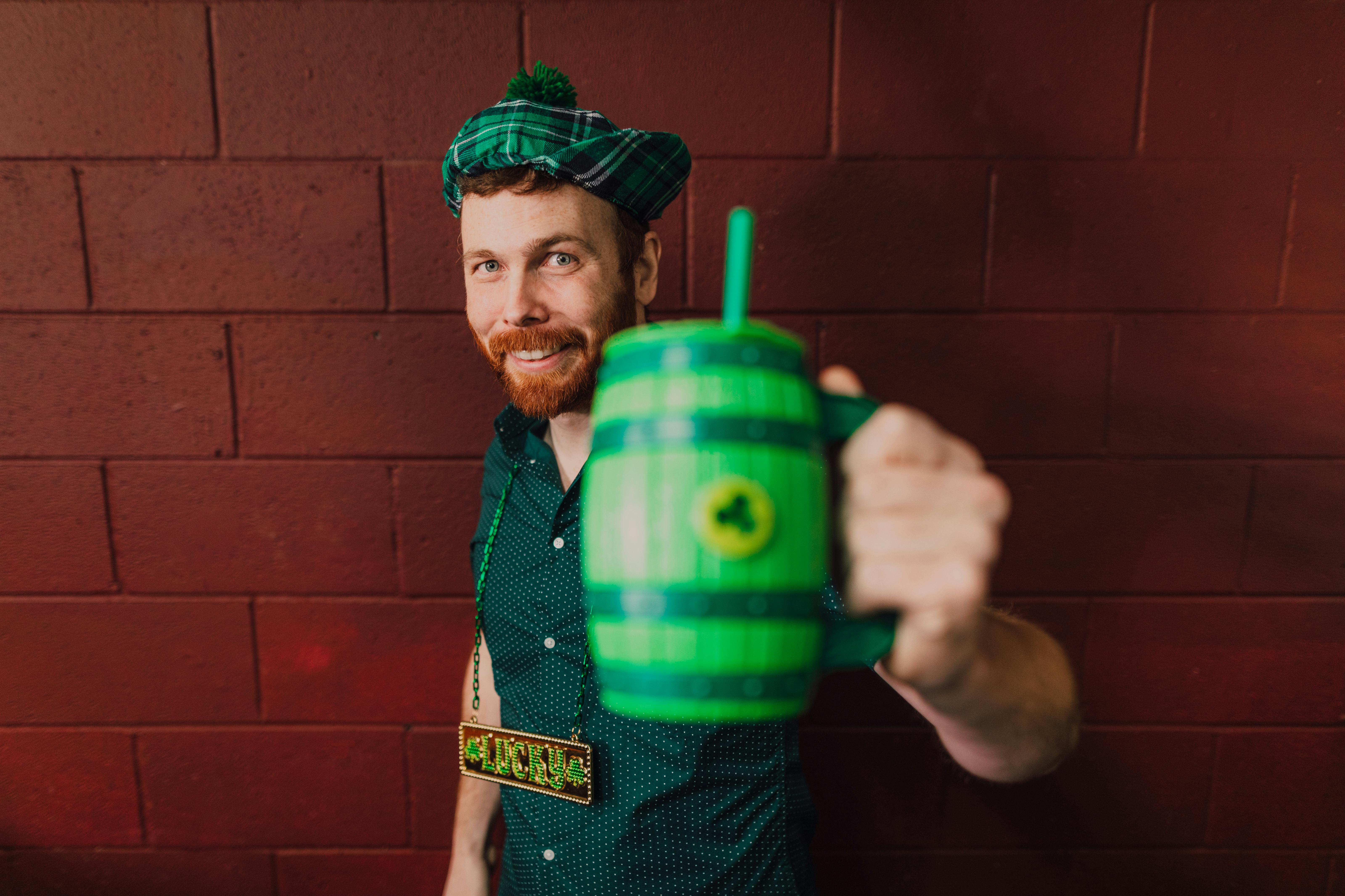 Happy man in Irish-themed outfit holding a green cup against red wall, celebrating Celtic culture.