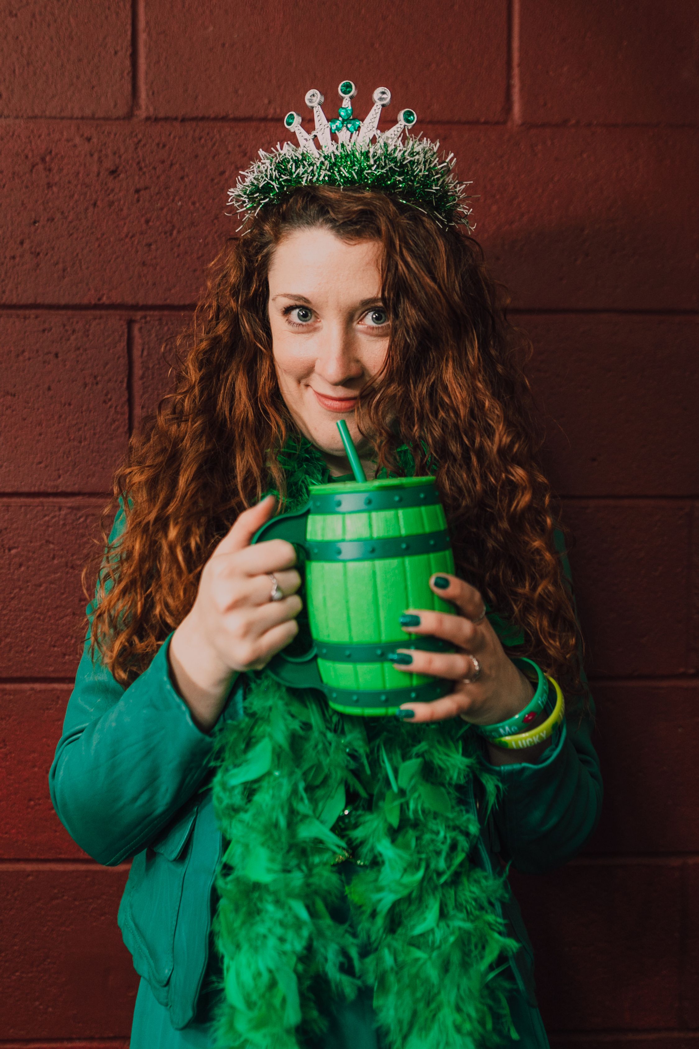 Smiling woman in festive green accessories holding a barrel mug against a red wall.