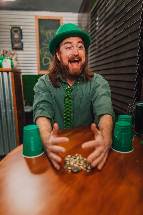 Free Joyful man in green attire celebrating St. Patrick's Day with gold coins and plastic cups on a wooden table. Stock Photo