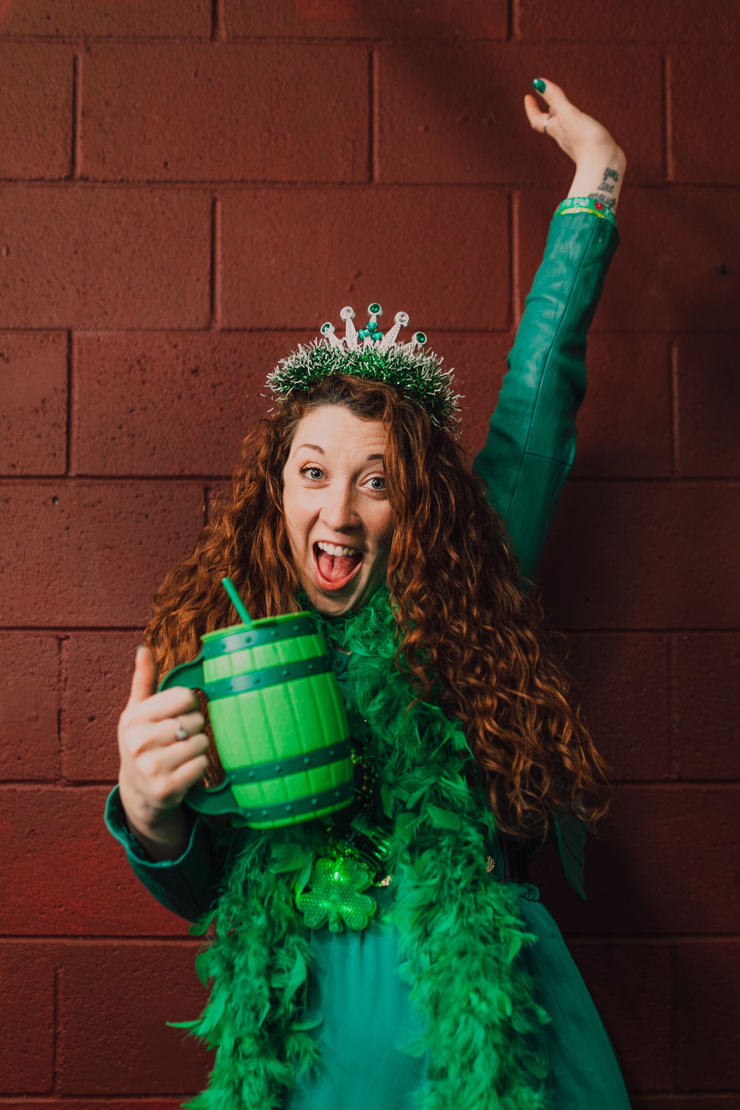 Cheerful woman in green attire celebrating St. Patrick's Day with a festive crown and mug.