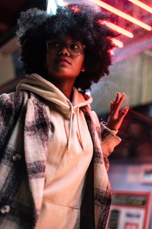 Free Serious African American young female in trendy coat and hoodie with eyeglasses standing near illuminated building window in city district at night while looking away Stock Photo