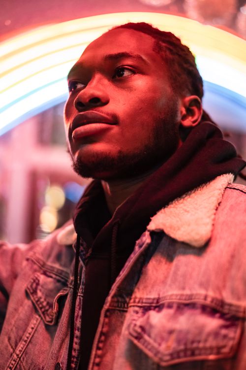 Free Pensive African American young man in trendy clothes near building with illuminated rainbow signboard at night in city district while looking away Stock Photo