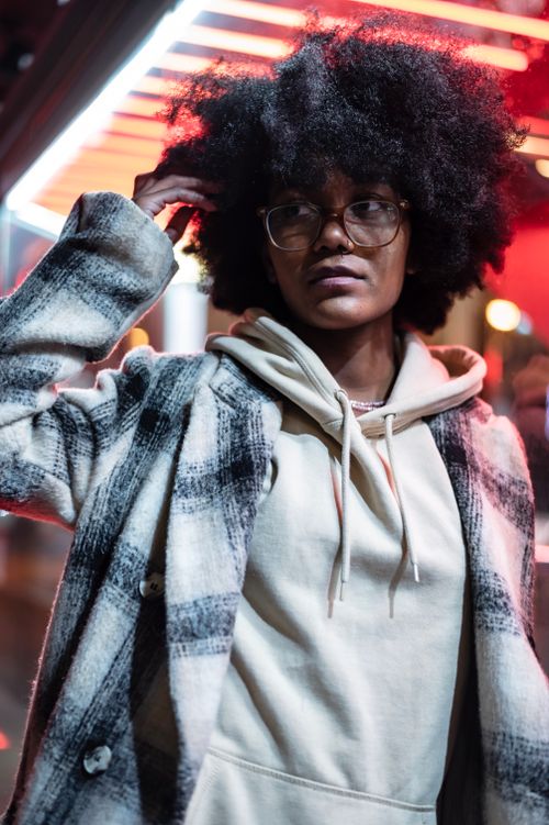 Free Pensive young black woman in hoodie and coat with eyeglasses standing on city street near glowing building at night while looking away Stock Photo