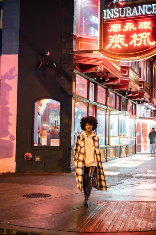 Full body of African American female in stylish casual wear walking on paved street with illuminated signboard and buildings