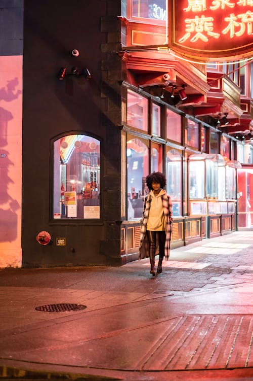 Black woman walking on sidewalk near illuminated buildings