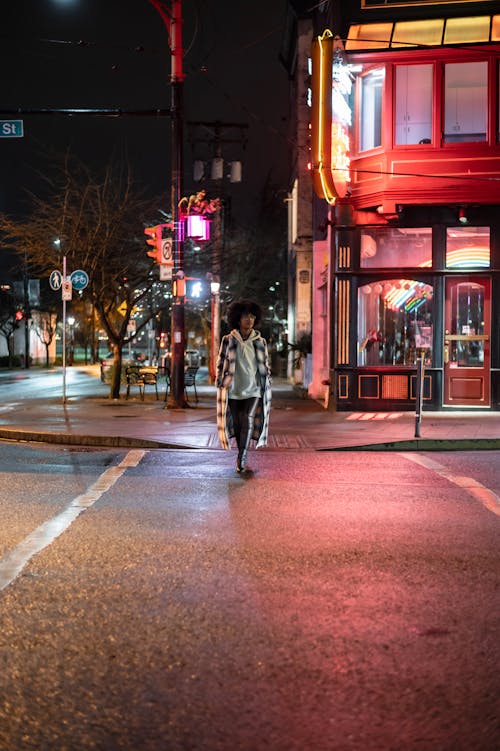 Full body of African American female with Afro hairstyle walking on pedestrian crossing against buildings with lights