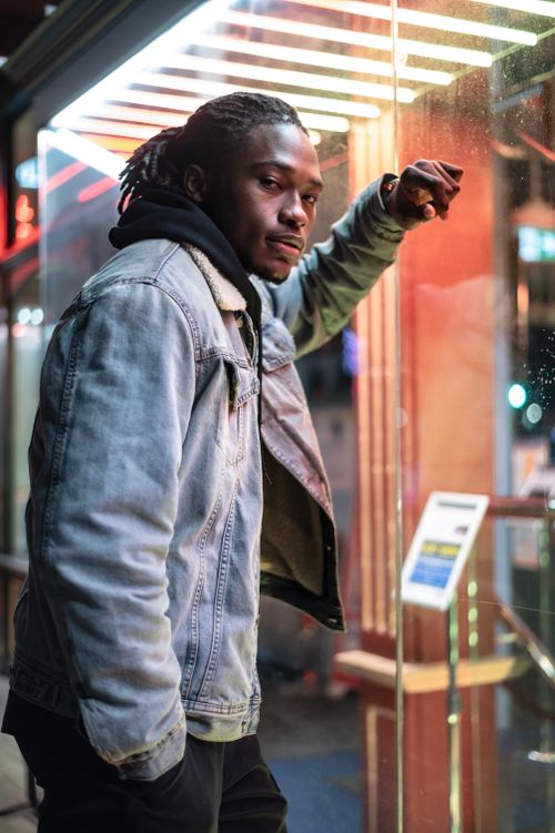 Free Serious African American male with dreadlocks looking at camera while standing near glass wall of modern glowing building with turnstile on dark street Stock Photo