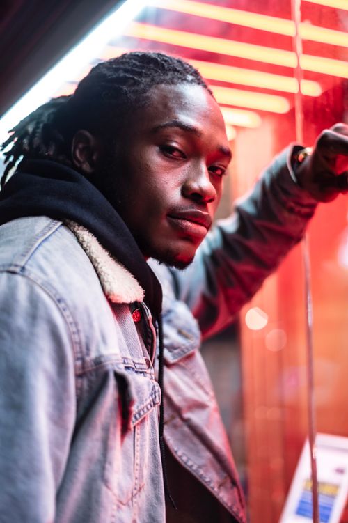 Free Serious African American male with dreadlocks wearing denim jacket looking at camera while standing near glass wall of modern building with glowing lights Stock Photo