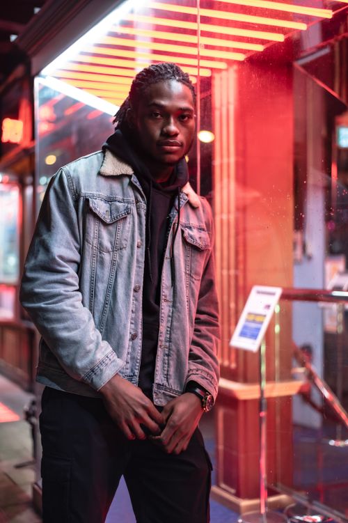 Free Confident African American male with dreadlocks and stylish denim jacket looking at camera while standing near glowing transparent building with turnstile on evening street Stock Photo