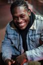 Happy African American male with dreadlocks wearing stylish denim jacket smiling and looking at camera on sidewalk against blurred background