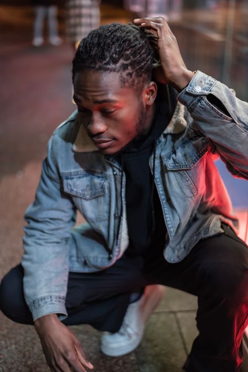 Free Stylish African American male wearing sneakers and denim jacket touching hair while sitting on haunches near building on evening street Stock Photo