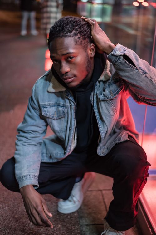 Free Serious African American male touching dreadlocks wearing denim jacket looking at camera while sitting on haunches near building in city Stock Photo