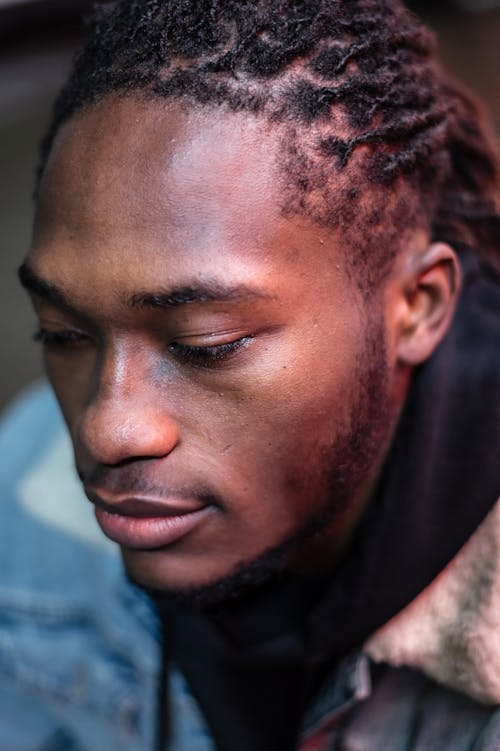 Calm African American man with dreadlocks and beard wearing denim jacket looking down while spending time on street in city